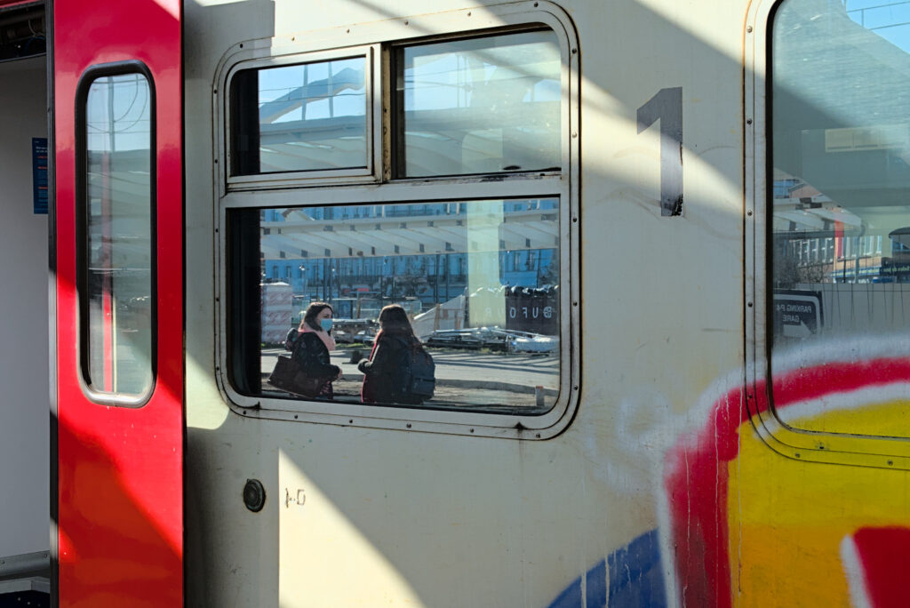 Vitre extérieure sur laquelle se reflètent deux jeune femmes qui discutent. On voit sur le train une porte rouge ouverte, une partie d'un tag rouge et jaune et le chiffre 1 en noir sur un fon gris.