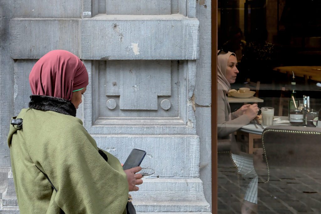 a woman in a hijab looking at her cell phone. Anothe woman is eating in a restaurant.