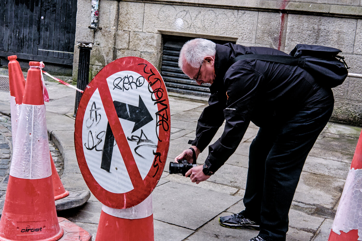 A man in black bends down with a camera to photograph a vandalized traffic sign on a pedestrian alley, surrounded by traffic cones.